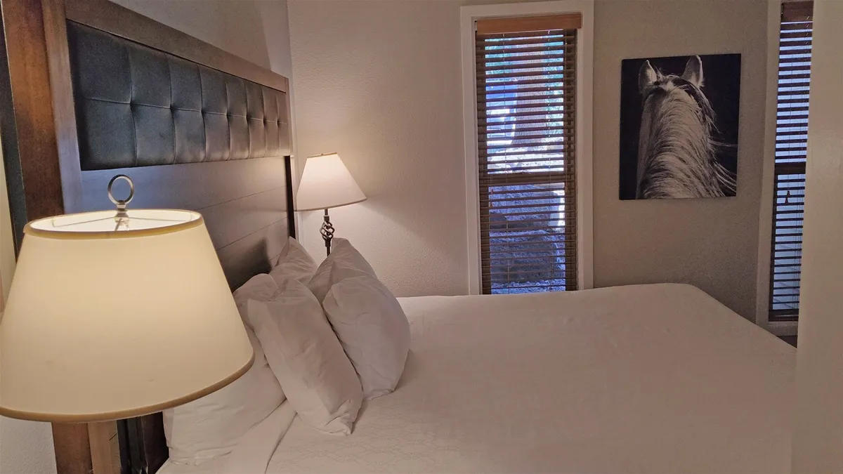 A serene king bedroom featuring a bed with crisp white linens, a warm lampshade on the nightstand, striking horse artwork above the headboard, and a window with blinds that lets in gentle natural light. The overall ambiance is tranquil and inviting, perfect for restful nights.