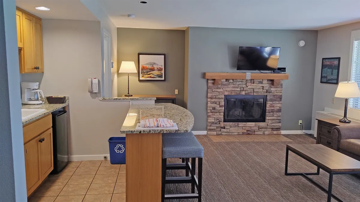 The image showcases a warmly lit, inviting condo interior at Seventh Mountain Resort. The focal points include a modern kitchen area, a cozy fireplace, a flat-screen television, and a comfortable seating space, all arranged for relaxation and socializing. The ambiance is perfect for unwinding after outdoor adventures, with soft lighting and stylish decor.