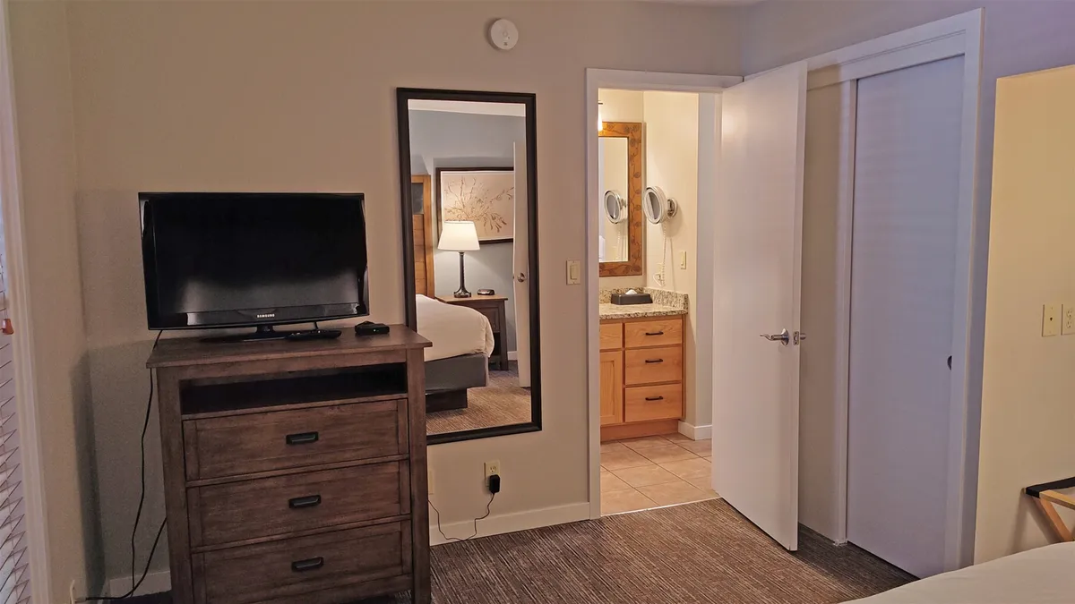 A cozy bedroom angle featuring a dresser, mirror, television, and the bathroom door visible. The scene emphasizes the direct connection between the bedroom and the bathroom, underlining privacy and easy access.