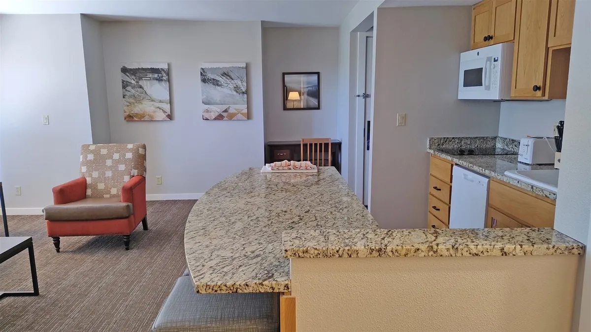A welcoming kitchen scene featuring a granite countertop, red accent chair, and visible kitchen appliances. The countertop is spacious, with ample room for meal prep or casual dining, and the presence of dishware and cookware hints at easy home cooking. Artistic touches on the wall add to the inviting, homey atmosphere.