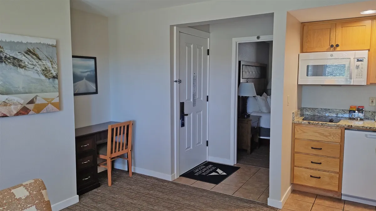 An inviting interior view highlighting a dedicated desk and chair set against a wall adorned with nature-inspired artwork. Nearby, a kitchenette and the doorway to a bedroom suggest an open-concept design ideal for both productivity and relaxation.