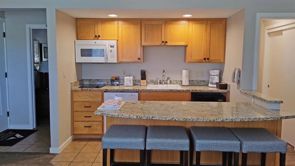 A kitchen scene featuring a granite countertop, kitchen island, wooden cabinets, and bar stools. The setup suggests both meal prep and casual dining are easy and enjoyable, with plenty of space to gather and interact. Warm lighting and practical design contribute to a comfortable, functional environment.