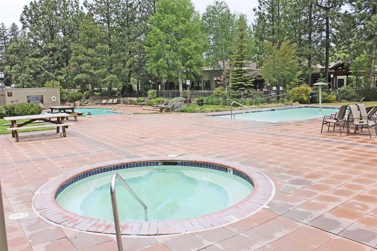 This inviting exterior image features a sparkling swimming pool surrounded by lush green trees, a relaxing hot tub, and picnic tables. The scene is vibrant with touches of activity, showcasing spaces for swimming, relaxing in the hot tub, or gathering for picnics amidst tranquil natural surroundings.