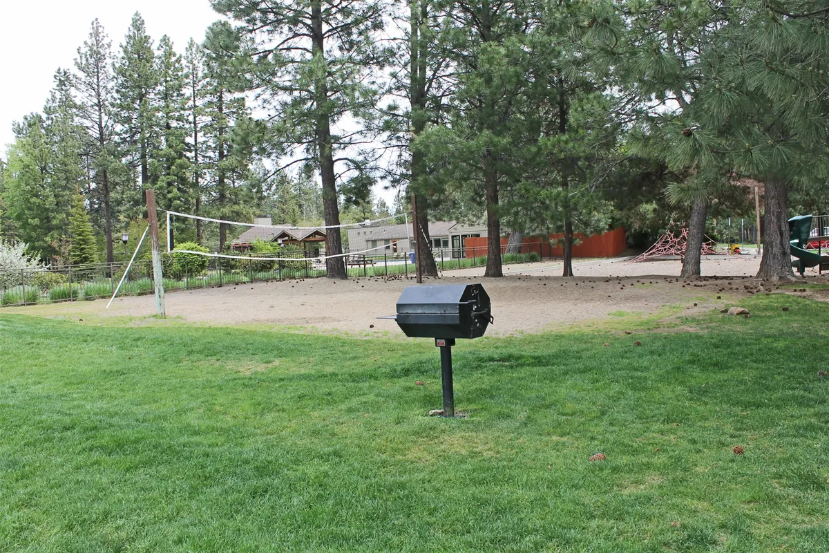 This image showcases an outdoor volleyball court set amidst lush green grass and leafy trees, with a barbecue grill nearby. The scene hints at communal fun and relaxation, with plenty of space for guests to gather, play, and enjoy the outdoors together.