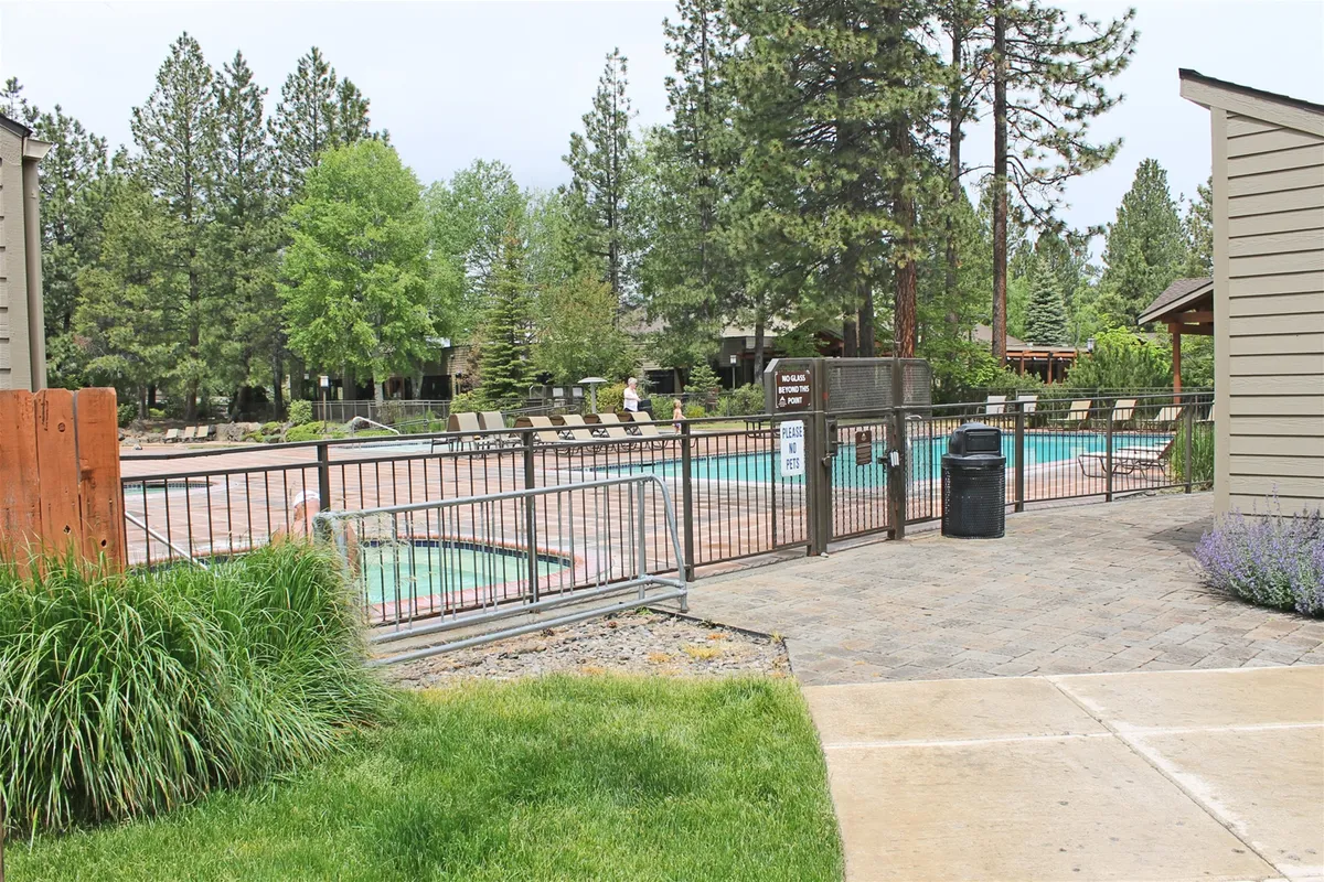 A sparkling swimming pool is surrounded by lounge chairs, grassy areas, and mature trees, enclosed by a fence for privacy and safety. The space radiates relaxation and community, ideal for both quiet moments and social gatherings.