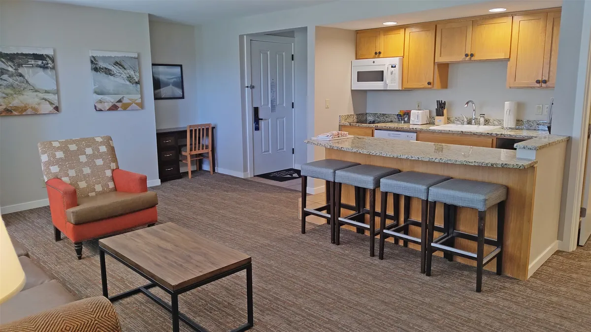 A wide-angle view capturing the kitchen area, breakfast bar with bar stools for four, a red accent chair, and stylish wall art. The open layout seamlessly connects the kitchen to the living space, emphasizing sociability and easy interaction while cooking or dining. The breakfast bar is a focal point, ideal for casual meals or conversation.
